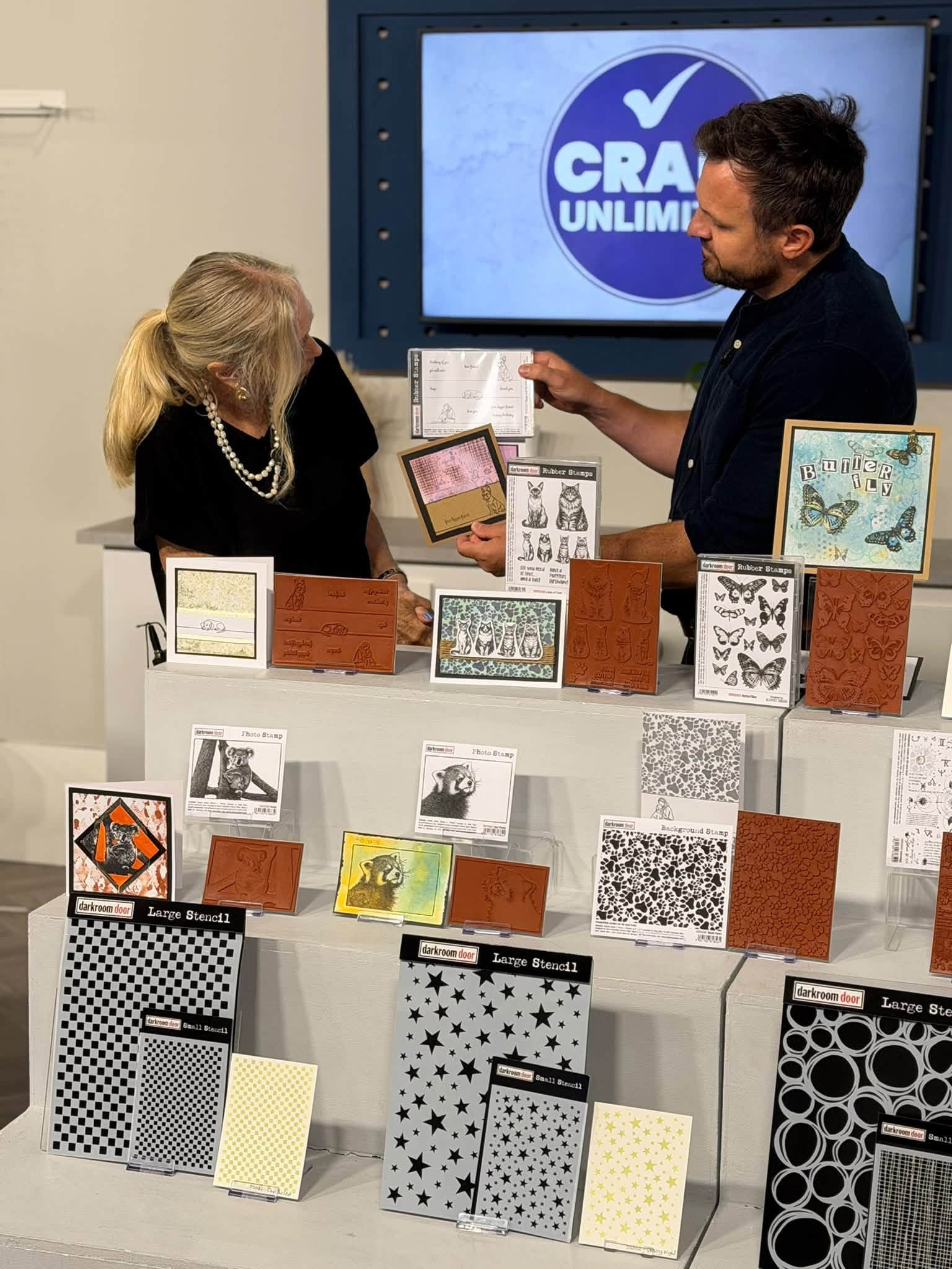 The presenter holding a stamp set beside Karen, with stamps, stencils and sample cards arranged in front